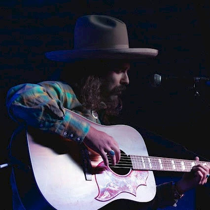 A bearded musician wearing a wide-brim hat plays an acoustic guitar on stage.