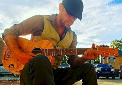 A man wearing a cap and vest sits on a rock playing an acoustic guitar outdoors, with a blue sky and parked cars in the background.