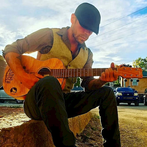 A man wearing a cap and vest sits on a rock playing an acoustic guitar outdoors, with a blue sky and parked cars in the background.