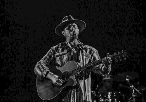A guitarist wearing a hat and plaid shirt plays an acoustic guitar on stage, singing into a microphone, with a dark background.
