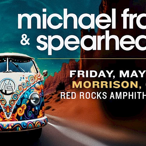 A colorful vintage VW bus drives on a desert road toward a concert poster for Michael Franti & Spearhead, showing date and location.
