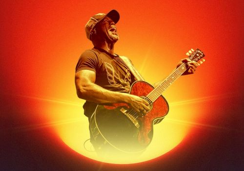 A guitarist wearing a cap plays an acoustic guitar against a dramatic orange-red sunset backdrop, glowing light radiating behind him.