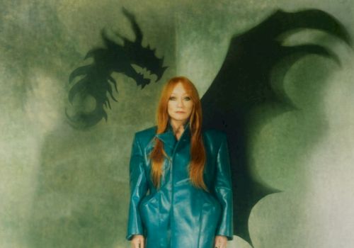 A woman with long red hair in a teal coat stands before a green background with dragon silhouettes behind her, creating a fantasy vibe.