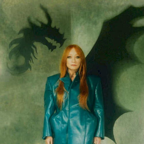 A woman with long red hair in a teal coat stands before a green background with dragon silhouettes behind her, creating a fantasy vibe.