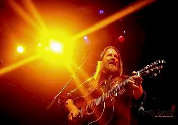 A guitarist performing on stage under bright yellow and orange lights, long hair, singing into a microphone, energetic live concert vibe.