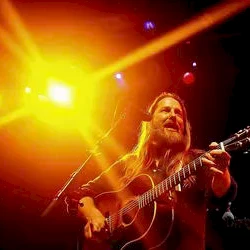 A guitarist performing on stage under bright yellow and orange lights, long hair, singing into a microphone, energetic live concert vibe.