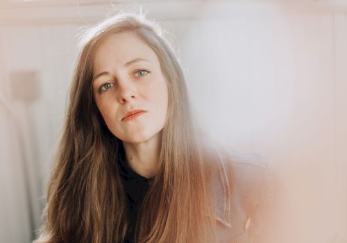 A young woman with long brown hair gazes softly at the camera, soft light creating a dreamy, hazy vibe around her.