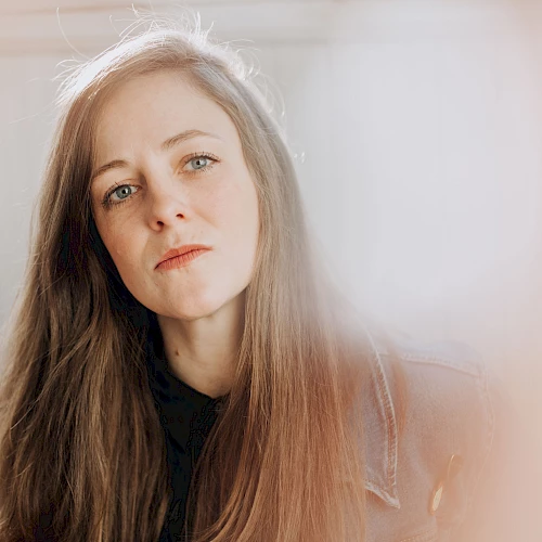 A young woman with long brown hair gazes softly at the camera, soft light creating a dreamy, hazy vibe around her.