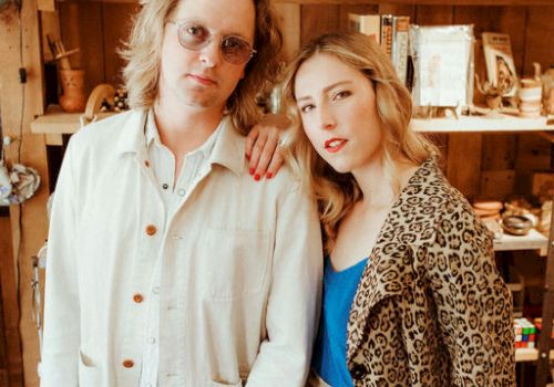 Two stylish friends pose in a cozy thrift-store setting, wearing bold patterns and soft smiles as they lean close for the shot.