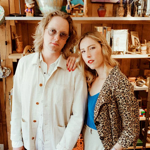 Two stylish friends pose in a cozy thrift-store setting, wearing bold patterns and soft smiles as they lean close for the shot.