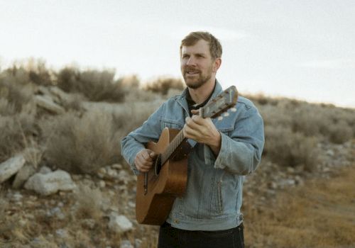 A man in a light denim jacket plays an acoustic guitar outdoors on a rocky, arid landscape, eyes closed, capturing a rustic, folk vibe.
