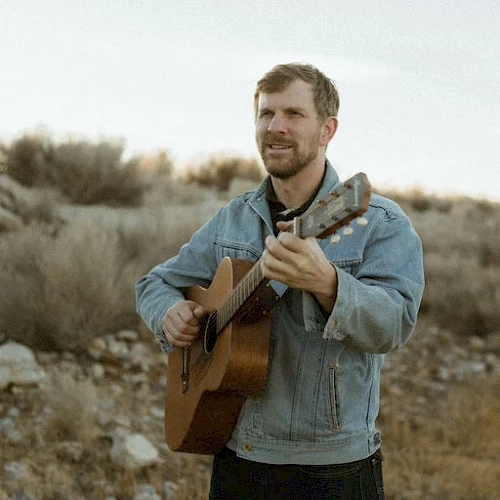 A man in a light denim jacket plays an acoustic guitar outdoors on a rocky, arid landscape, eyes closed, capturing a rustic, folk vibe.