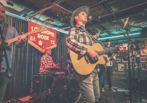 A live band performs on a cozy stage: guitarist singing, drummer, bassist, and another guitarist in plaid shirt under warm club lights.
