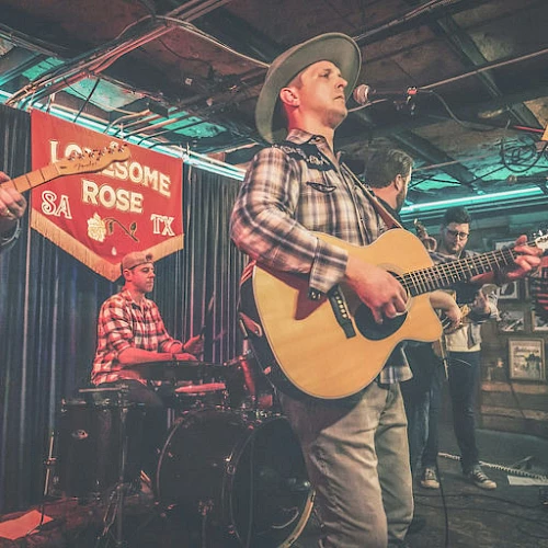 A live band performs on a cozy stage: guitarist singing, drummer, bassist, and another guitarist in plaid shirt under warm club lights.