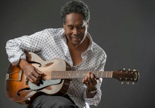 A man plays an acoustic guitar, seated, smiling, wearing a white patterned shirt against a dark gray backdrop.