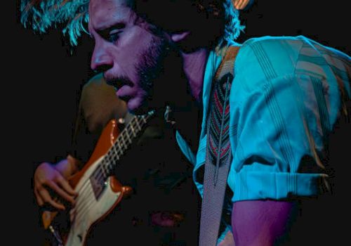 A musician playing an electric guitar on stage, blue lighting and a focused, energetic performance.