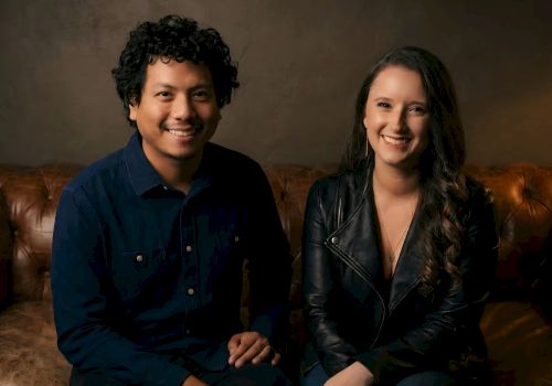 Two people sitting on a brown leather couch, smiling for the camera, a casual studio setting.