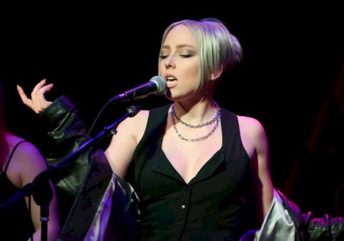 A female singer with short blonde hair performs on stage, wearing a black outfit and necklace, holding a microphone with a jacket draped.