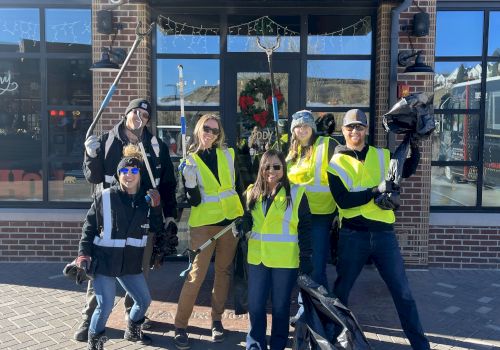 A group of eight people wearing yellow safety vests pose happily outside a storefront, some with raised arms, on a sunny day.