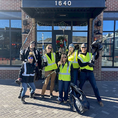 A group of eight people wearing yellow safety vests pose happily outside a storefront, some with raised arms, on a sunny day.