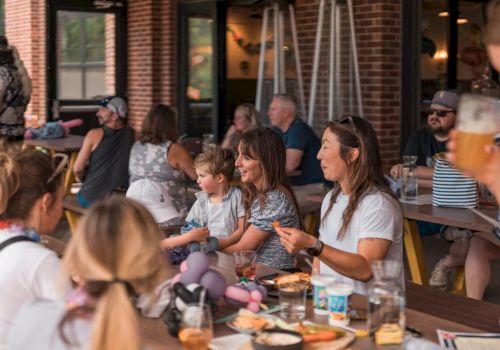 A group of people, adults and kids, sit around a long table at a sunny outdoor cafe, sharing food and drinks, smiling and talking.
