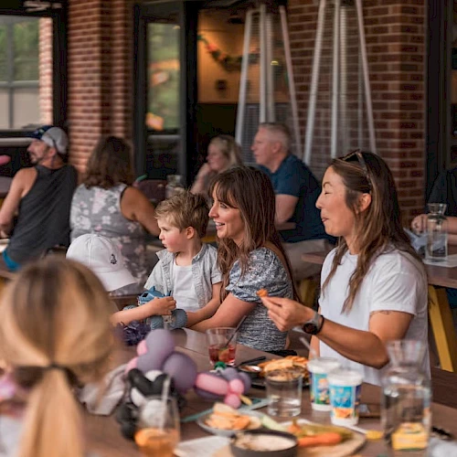 A group of people, adults and kids, sit around a long table at a sunny outdoor cafe, sharing food and drinks, smiling and talking.