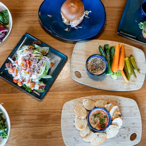 A table of salads, sandwiches and burgers with vegetable sticks, bread, and small cups of dressing/coffee for dipping.