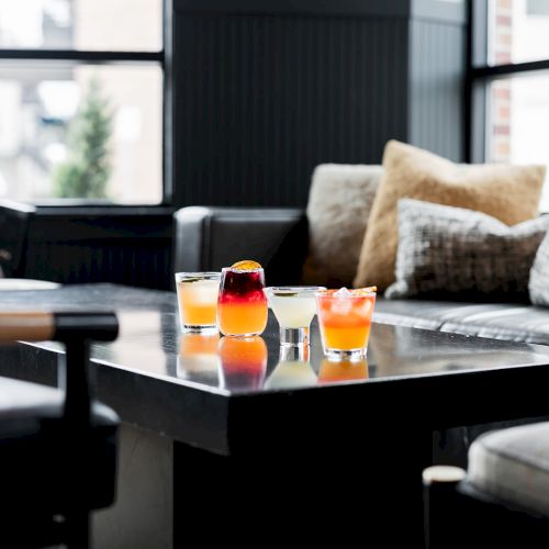 A modern living room with a glass coffee table holding colorful cocktails in shot glasses and a sofa with pillows.