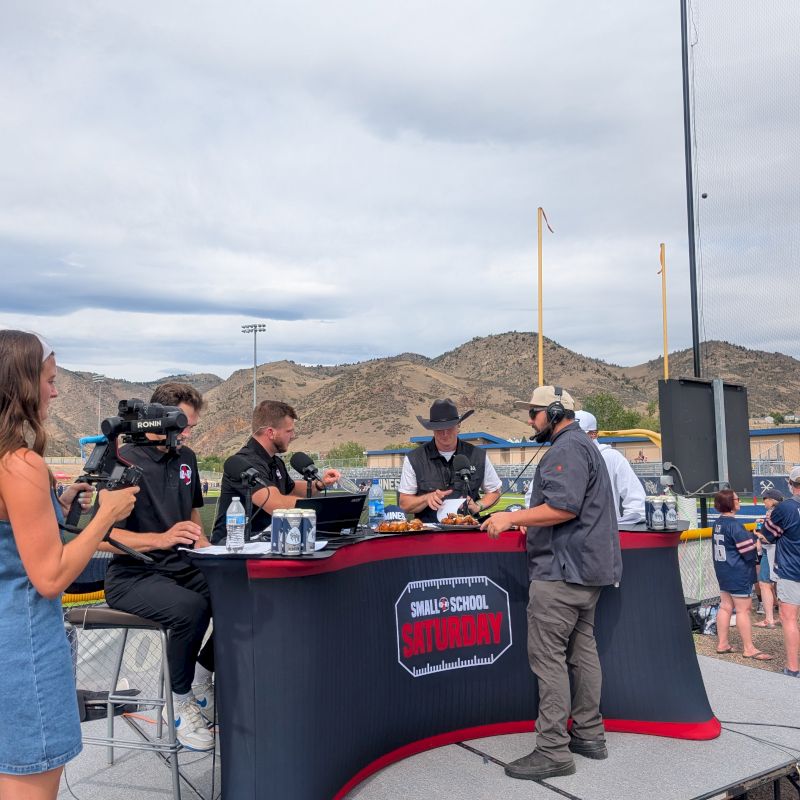 News crew records an interview at a sports field, with cameras and microphones, under cloudy skies and mountain hills behind them.