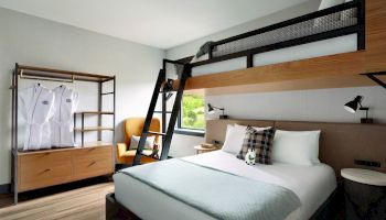 The image shows a modern hotel room with a double bed, an overhead bunk bed with a ladder, a wardrobe, and a yellow chair near a window.