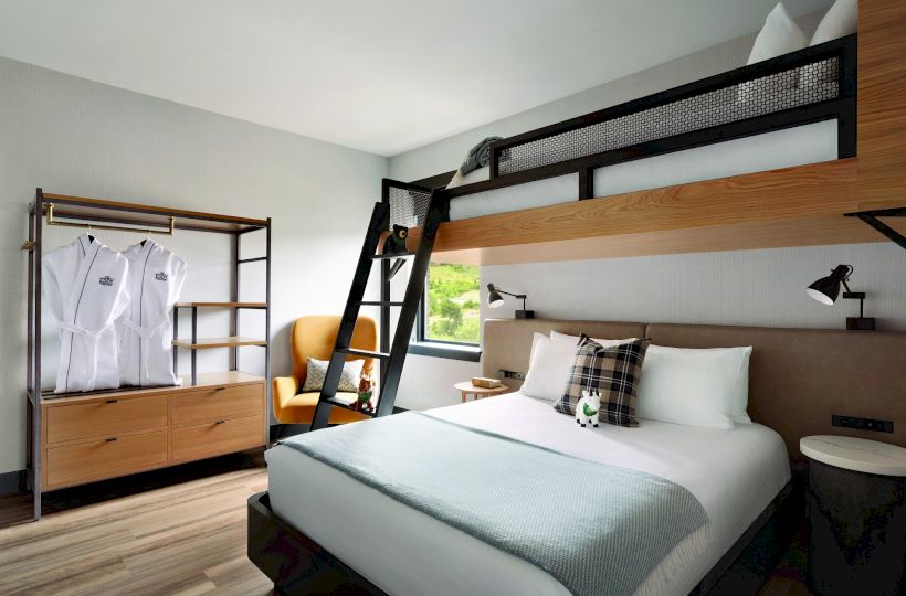 The image shows a modern hotel room with a double bed, an overhead bunk bed with a ladder, a wardrobe, and a yellow chair near a window.
