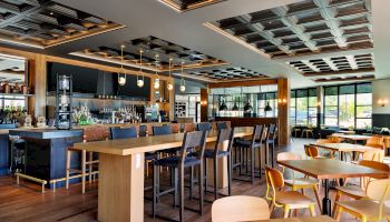 A modern restaurant interior with wooden furniture, a central bar area with high stools, ceiling lights, and large windows.