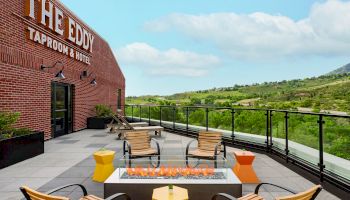 A rooftop patio with lounge chairs around a modern fire pit overlooks lush hills and greenery, beside a building labeled "The Eddy Taproom & Hotel."