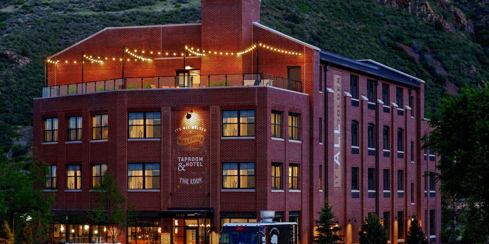 A multi-story brick building with outdoor string lights, labeled as a taproom and hotel, is situated near a grassy area and mountains.