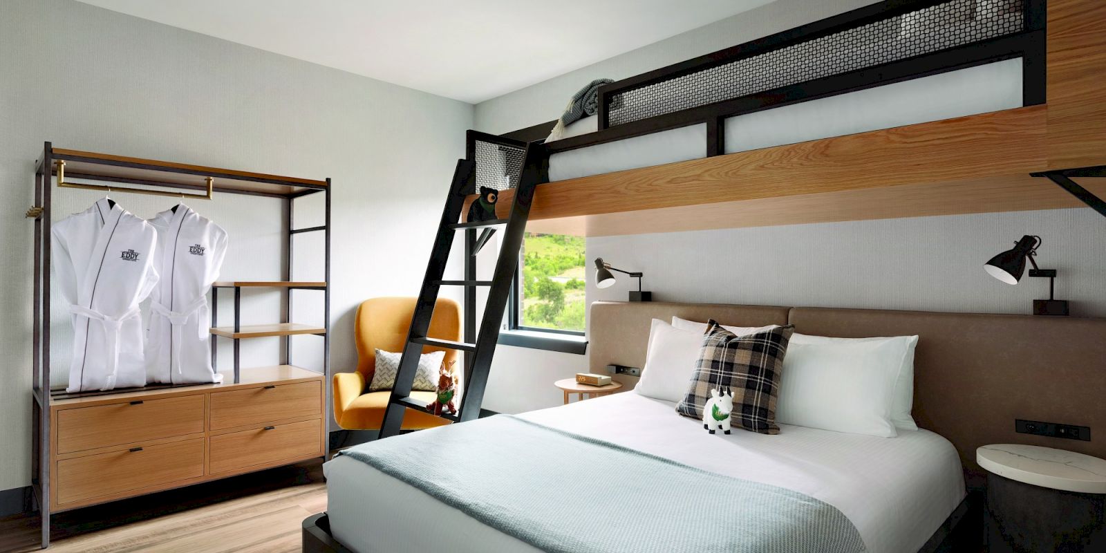 The image shows a modern hotel room with a double bed and a loft bed, a ladder, an armchair, a rack with robes, and wall-mounted reading lights.