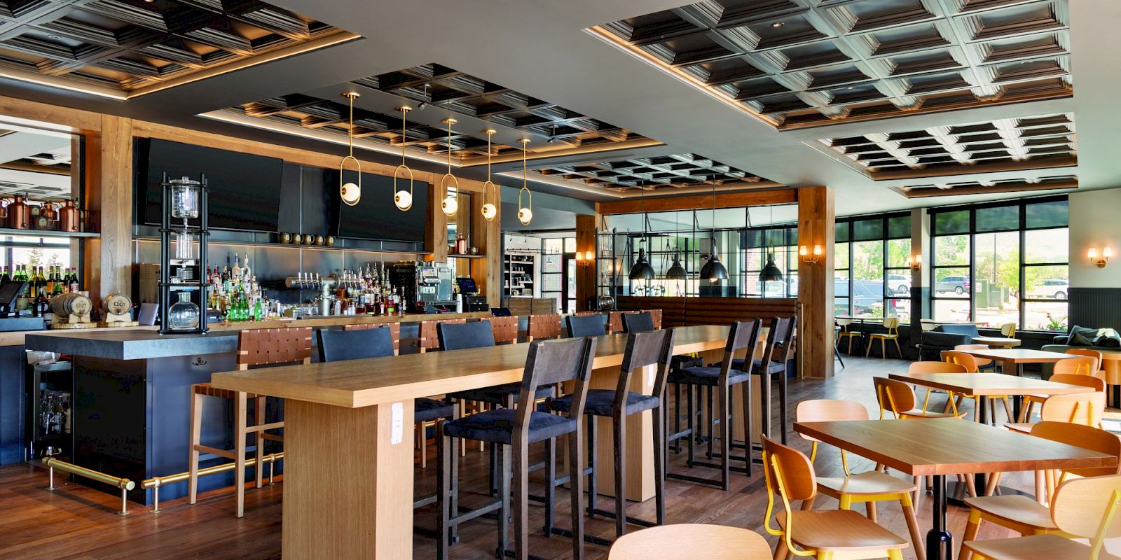 The image shows a modern, well-lit restaurant with wooden tables and chairs, a long bar with barstools, and stylish ceiling decor.