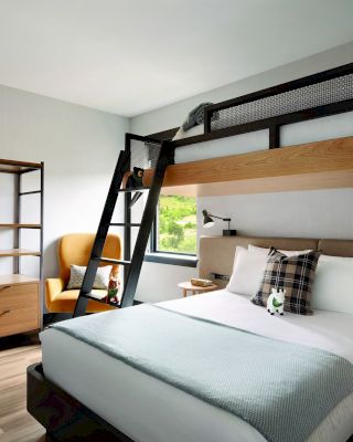 A modern hotel room with a double bed, an overhead bunk bed, a ladder, wardrobe, two bathrobes, an armchair, and a window offering natural light.