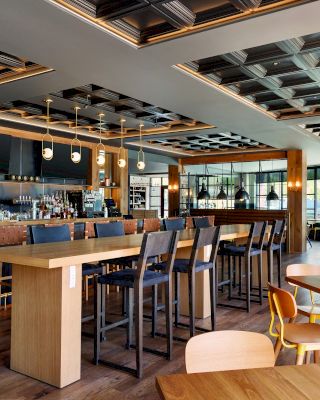 The image shows a modern, well-lit bar and restaurant with wooden tables, chairs, high stools, a well-stocked bar and contemporary decor.