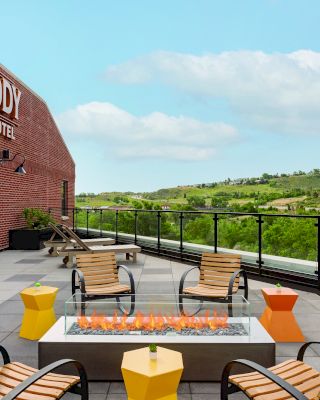 A rooftop patio with a fire pit, surrounded by chairs and tables, and a view of greenery. The sign reads "The Eddy Taproom & Hotel."