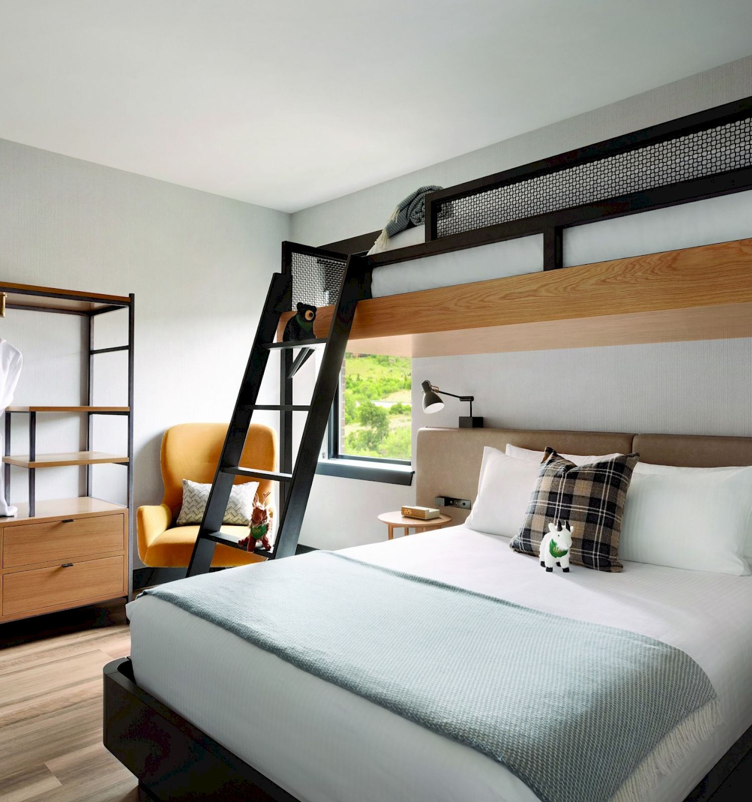 A modern hotel room with a double bed, an overhead bunk bed accessible by a ladder, and a wardrobe with two hanging robes.