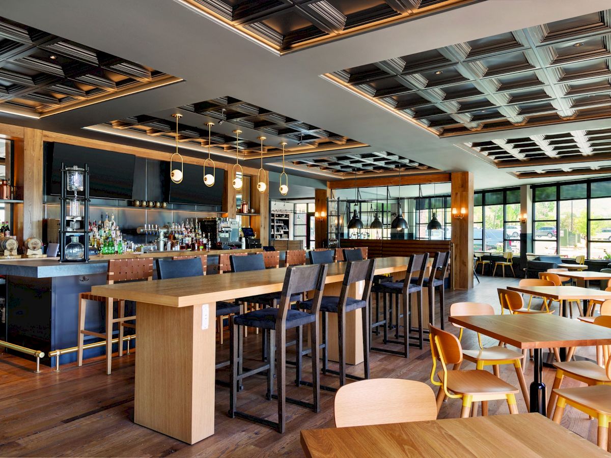 The image shows a modern, well-lit restaurant interior with wooden furniture, a bar area, and large windows providing natural light.