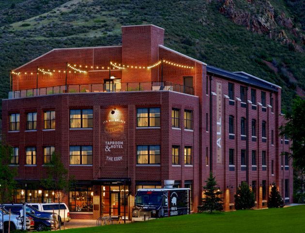 A charming multi-story brick building with string lights, surrounded by lush hills, features a brewery and hotel. Cars are parked in front.