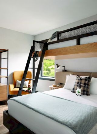 The image shows a modern hotel room with a double bed, a loft bed above accessed by a ladder, a wardrobe with robes, and a cozy chair by the window.
