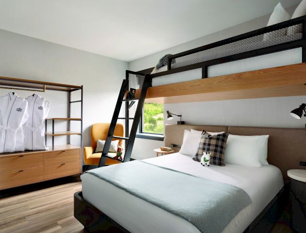 A modern hotel room with a double bed and a bunk bed, a small wardrobe with robes, an armchair, and a window.