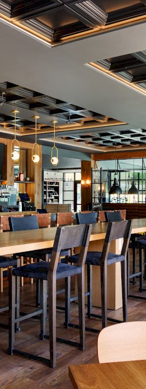 This image shows a modern restaurant interior with wooden furniture, high tables and stools, a bar area, and large windows allowing natural light to enter the space.