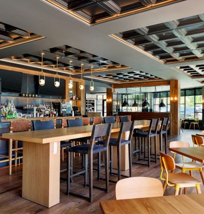 The image shows a modern restaurant interior with wooden tables and chairs, a bar counter with high stools, and a stylish ceiling design with recessed lighting.