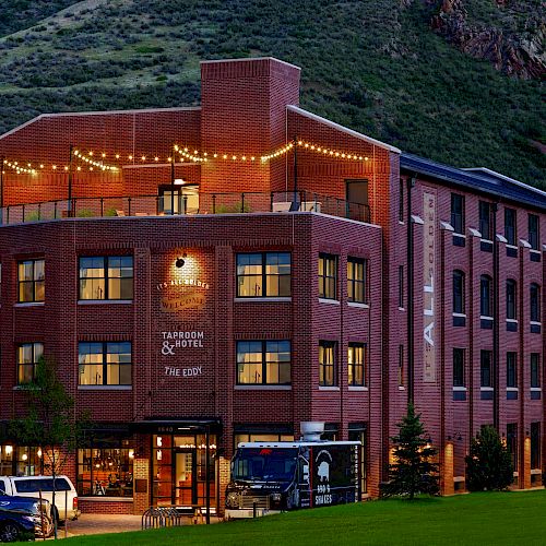 A brick building with string lights, signage for a taproom and hotel, and parked cars in front, set against a backdrop of green hills and trees.
