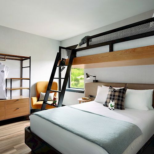 The image shows a modern bedroom with a bunk bed, a double bed below, a ladder, robes on a rack, an armchair, and a nightstand under a window.