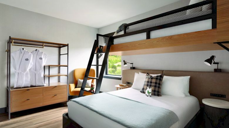 A modern hotel room features a bunk bed, a double bed, a ladder, a wardrobe with robes, a chair, and a window with a view of greenery, ending the sentence.