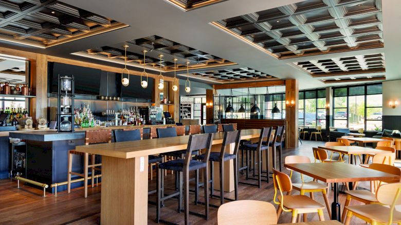 This image shows a modern restaurant interior with wooden furniture, a bar area with stools, shelves with drinks, and ceiling lights.